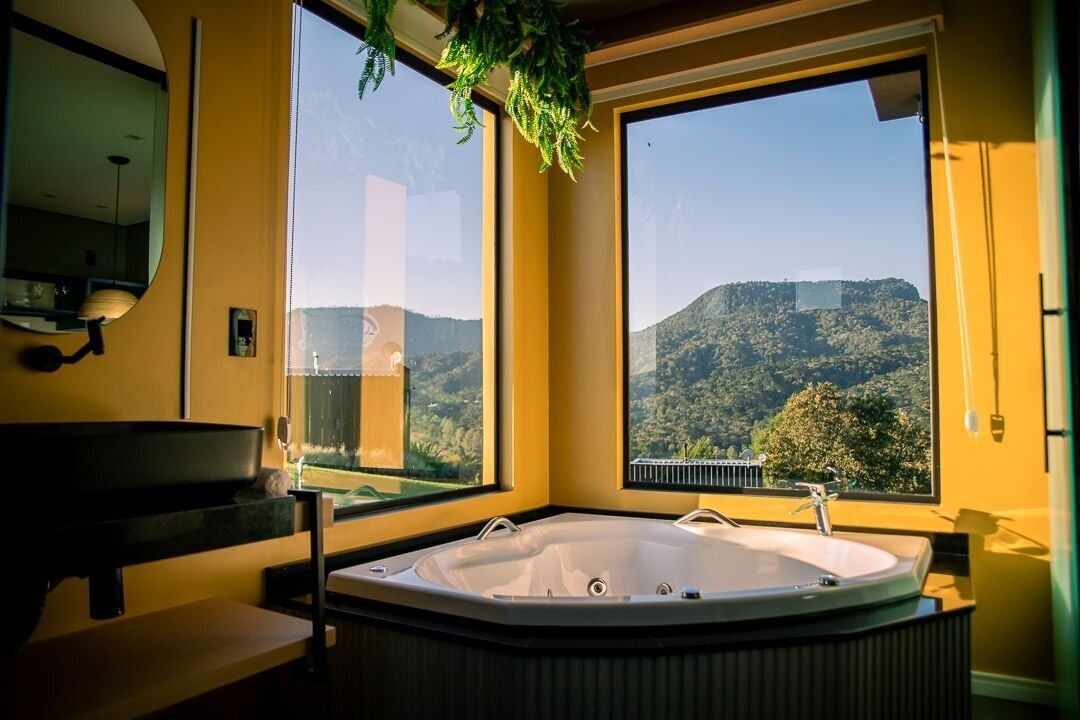 A bathroom with a large bathtub, a sink, and a mirror. There are two large windows offering a view of a mountainous landscape.