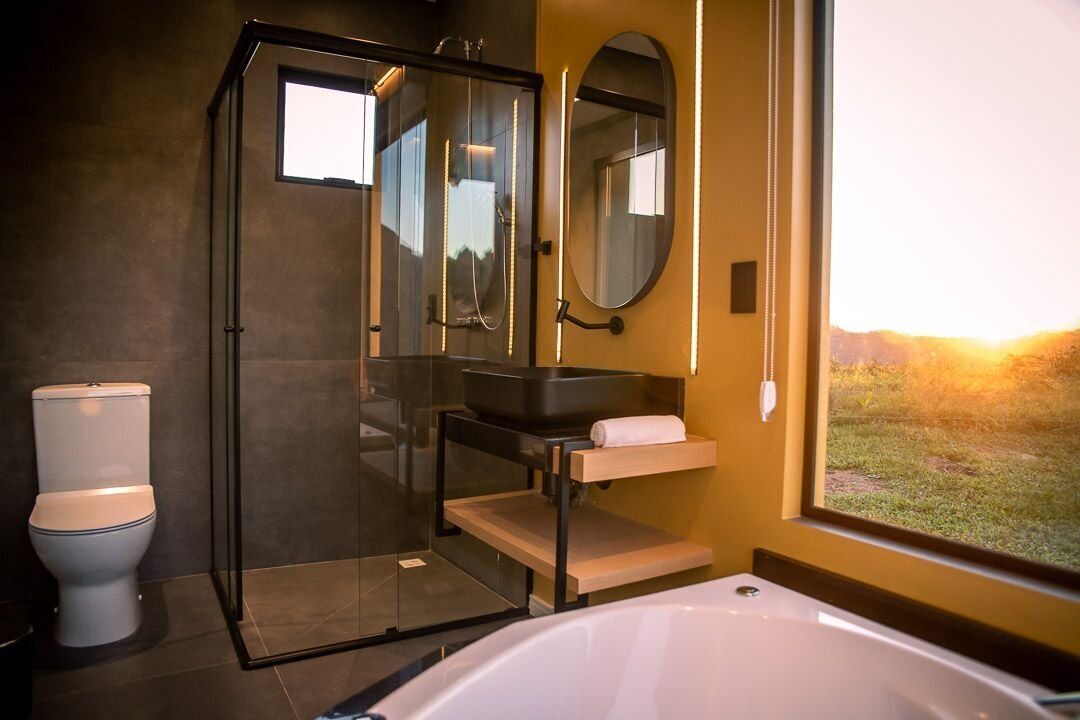 Un baño moderno con ducha acristalada, inodoro blanco, lavabo negro y una ventana grande con vistas a la puesta de sol.