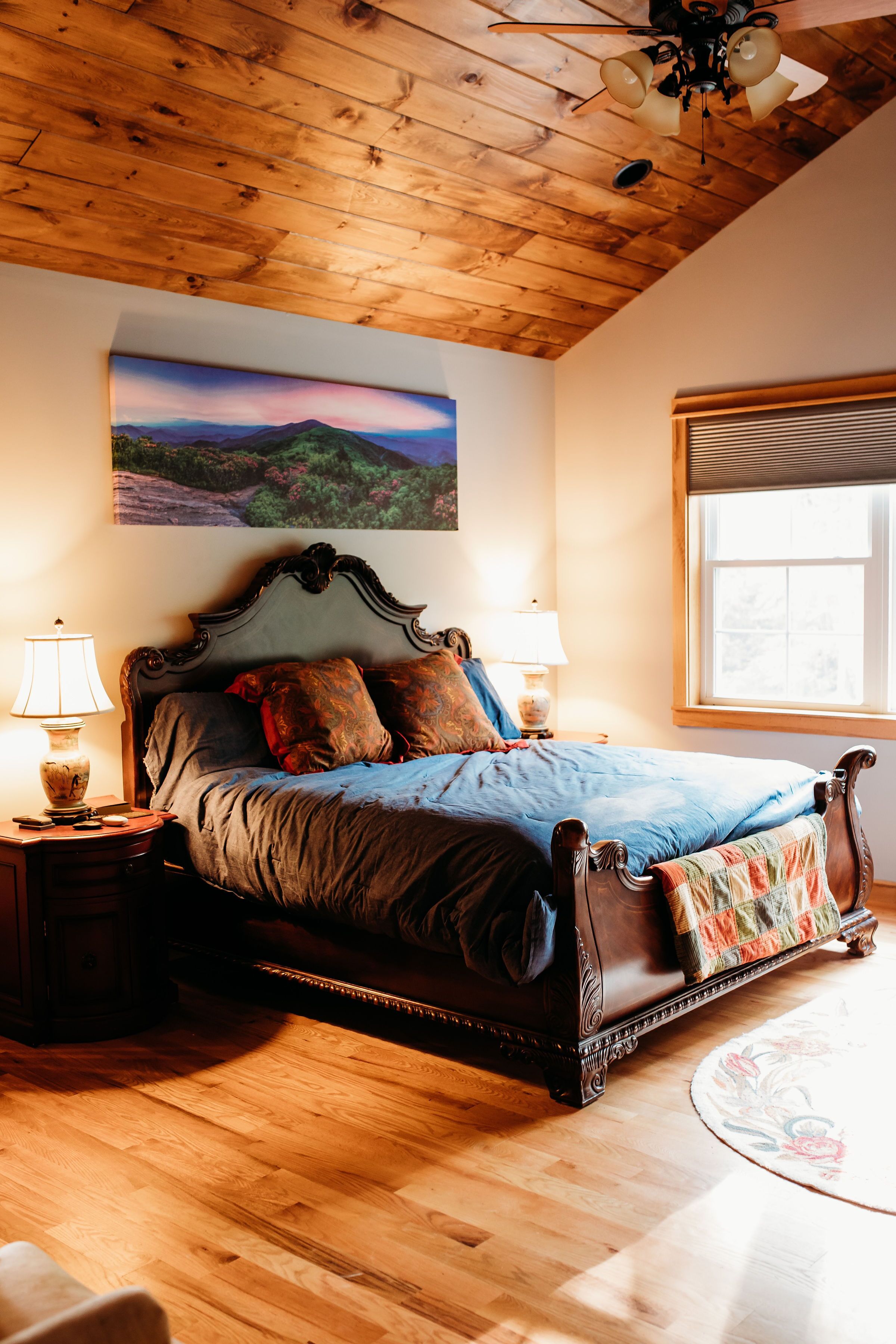 A bedroom with a wooden ceiling, a bed with a headboard, two bedside lamps, a window with blinds, and a painting on the wall.