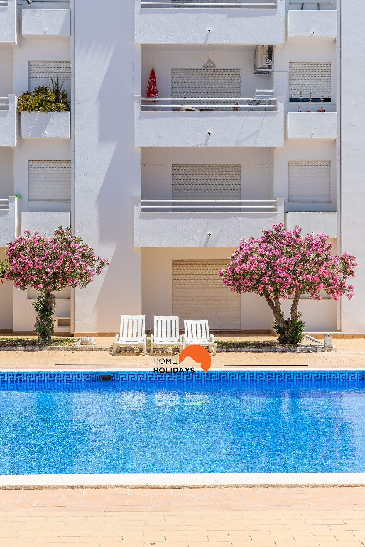 Ein modernes Hotel mit Schwimmbad, weißen Balkonen und zwei blühenden Bäumen.