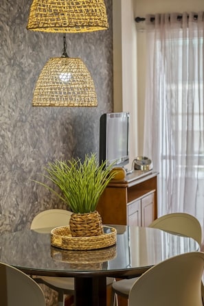 Un comedor con una mesa de vidrio, lámparas con pantalla tejida, una maceta tejida y un televisor sobre un mueble de madera.