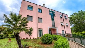 A pink building with multiple windows, a small entrance, and a paved pathway leading to a green fence. - Pink apartment 4 minutes walk from free beach (San Vincenzo)