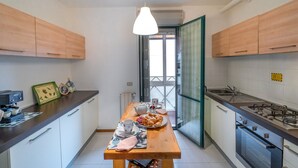 A narrow kitchen with wooden cabinets, a dining table set with plates, cups, and a teapot. There is a window with a view of the outside. - Pink apartment 4 minutes walk from free beach (San Vincenzo)