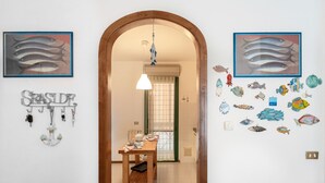A hallway with a wooden door leading to a dining area. There are framed fish art on the walls, a key rack, and a small table with a chair. - Pink apartment 4 minutes walk from free beach (San Vincenzo)