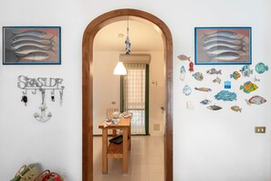 Un corridoio con una porta in legno che dà su un'area pranzo. Alle pareti sono presenti quadri con soggetti ittici, un portachiavi e un piccolo tavolo con una sedia.