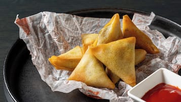 Una bandeja de samosas doradas sobre papel arrugado, acompañadas de un recipiente pequeño con salsa roja para mojar.