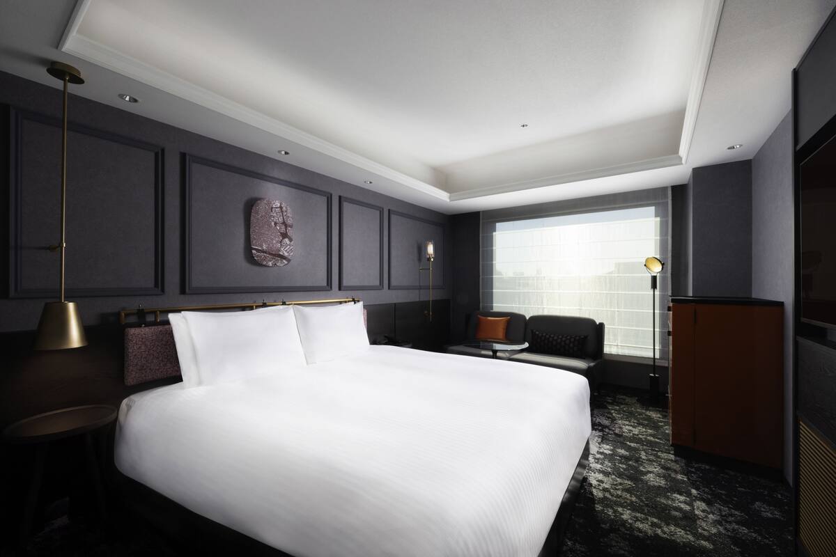 Habitación de hotel moderna con una cama grande, zona de estar con sofá y sillón, escritorio y un ventanal con vistas a la ciudad.