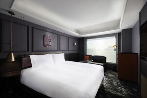 A modern hotel room with a large bed, a seating area with a sofa and armchair, a desk, and a large window offering a city view.