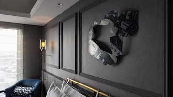 A modern hotel room with a large window, a blue armchair, a decorative wall piece, and a bedside table with a lamp.