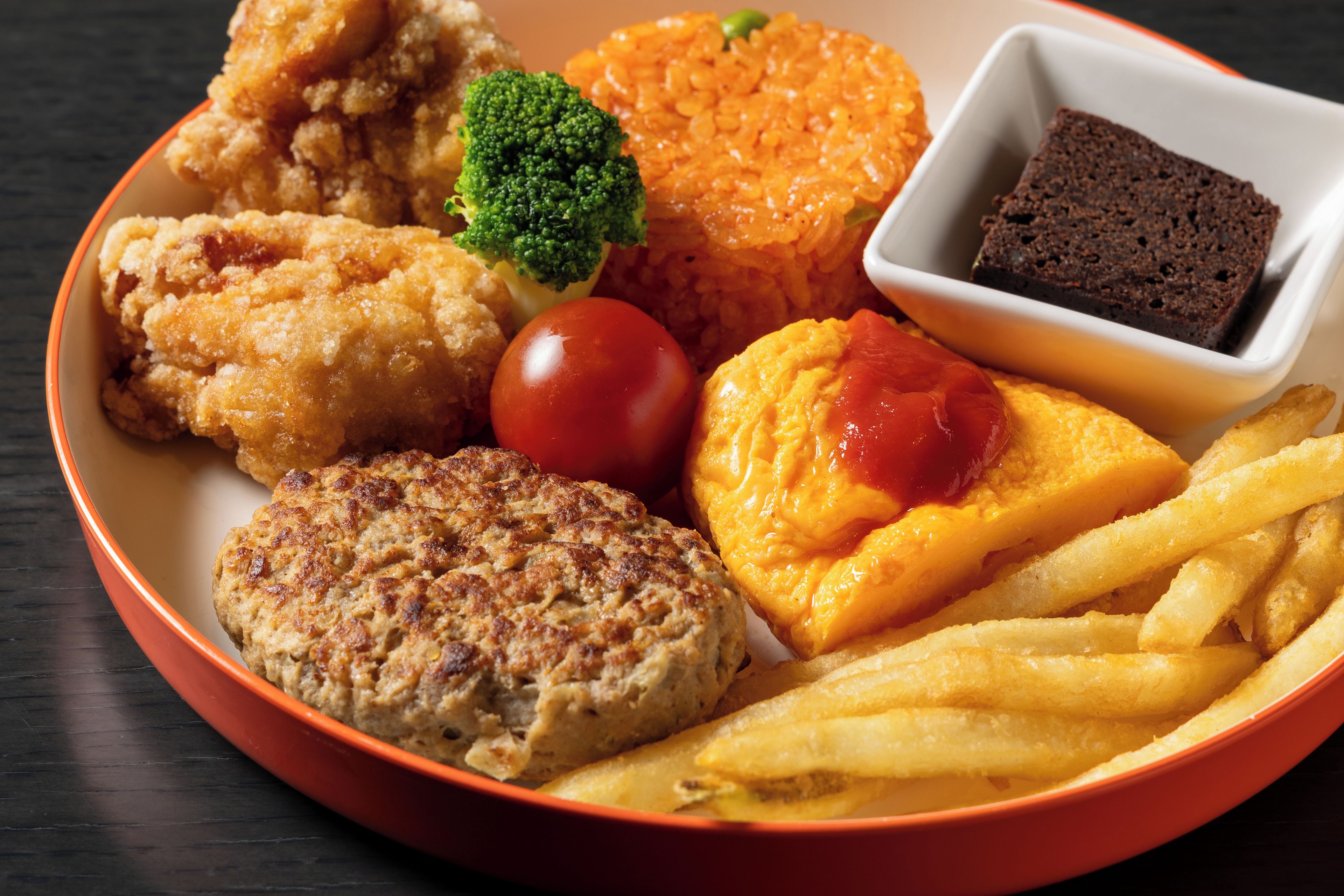 Un plato de comida con pollo frito, una hamburguesa, papas fritas, tomate, una rebanada de queso con kétchup, pan y un pequeño tazón de pastel de chocolate.