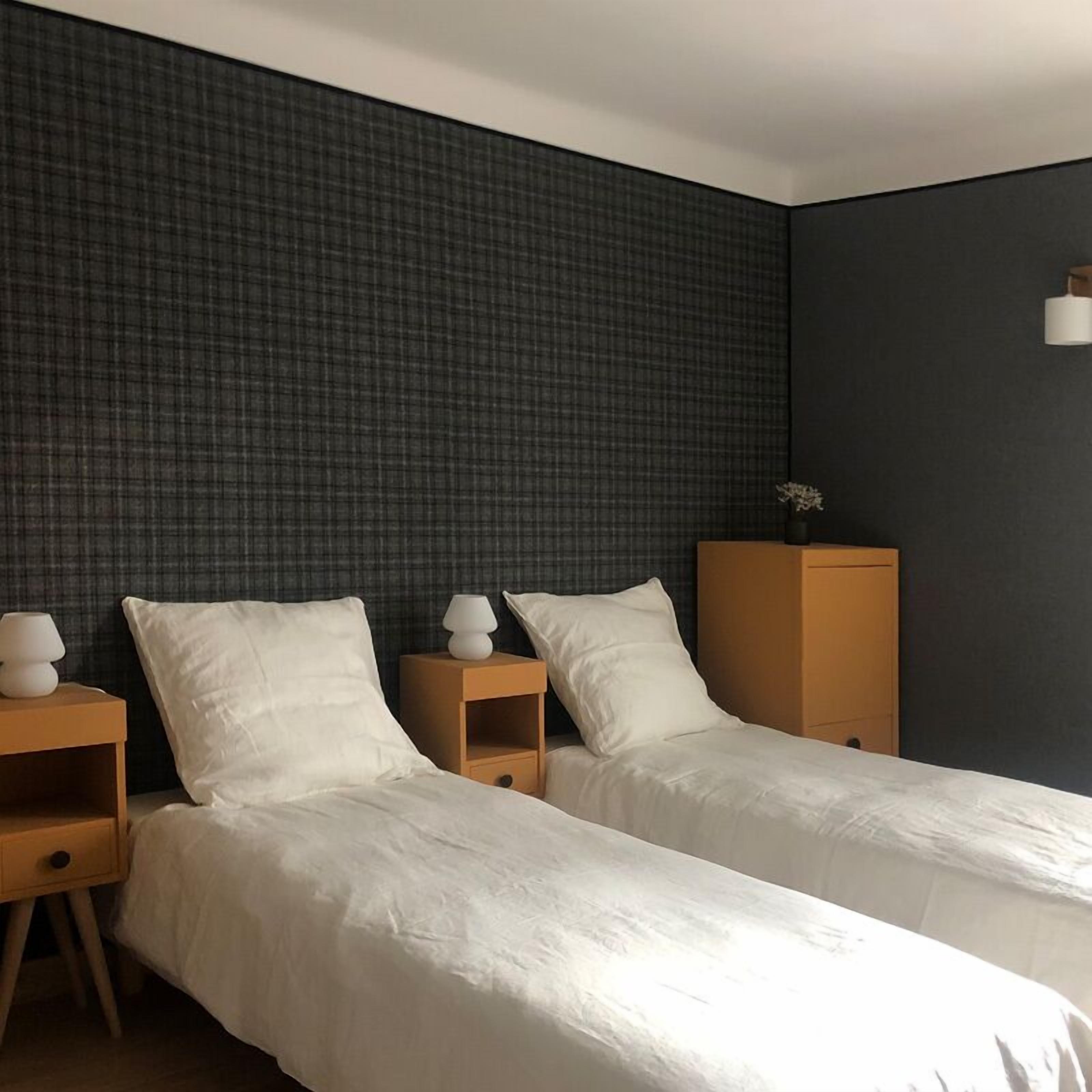 A hotel room with two single beds, a nightstand with a lamp, a wooden cabinet, and a dark textured wall.