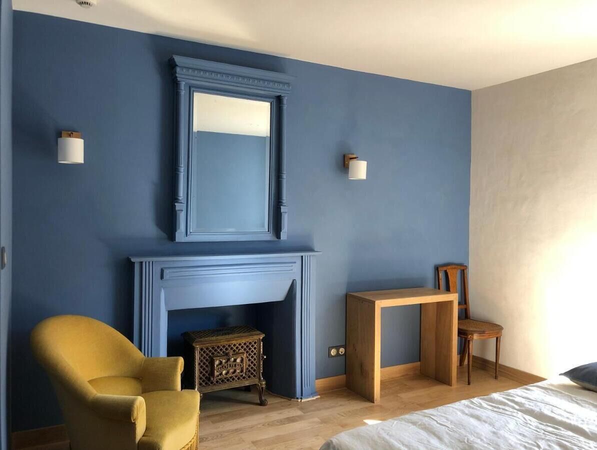 A room with a blue wall, a fireplace, a yellow armchair, a wooden table, and a wooden chair.