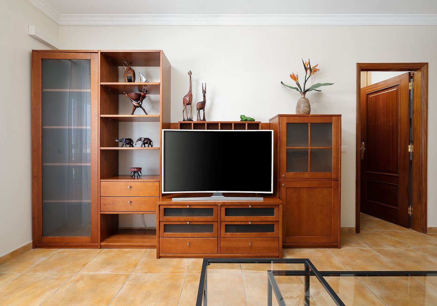 A living room with a TV on a wooden stand, a cabinet with glass doors, and a door leading to another room.