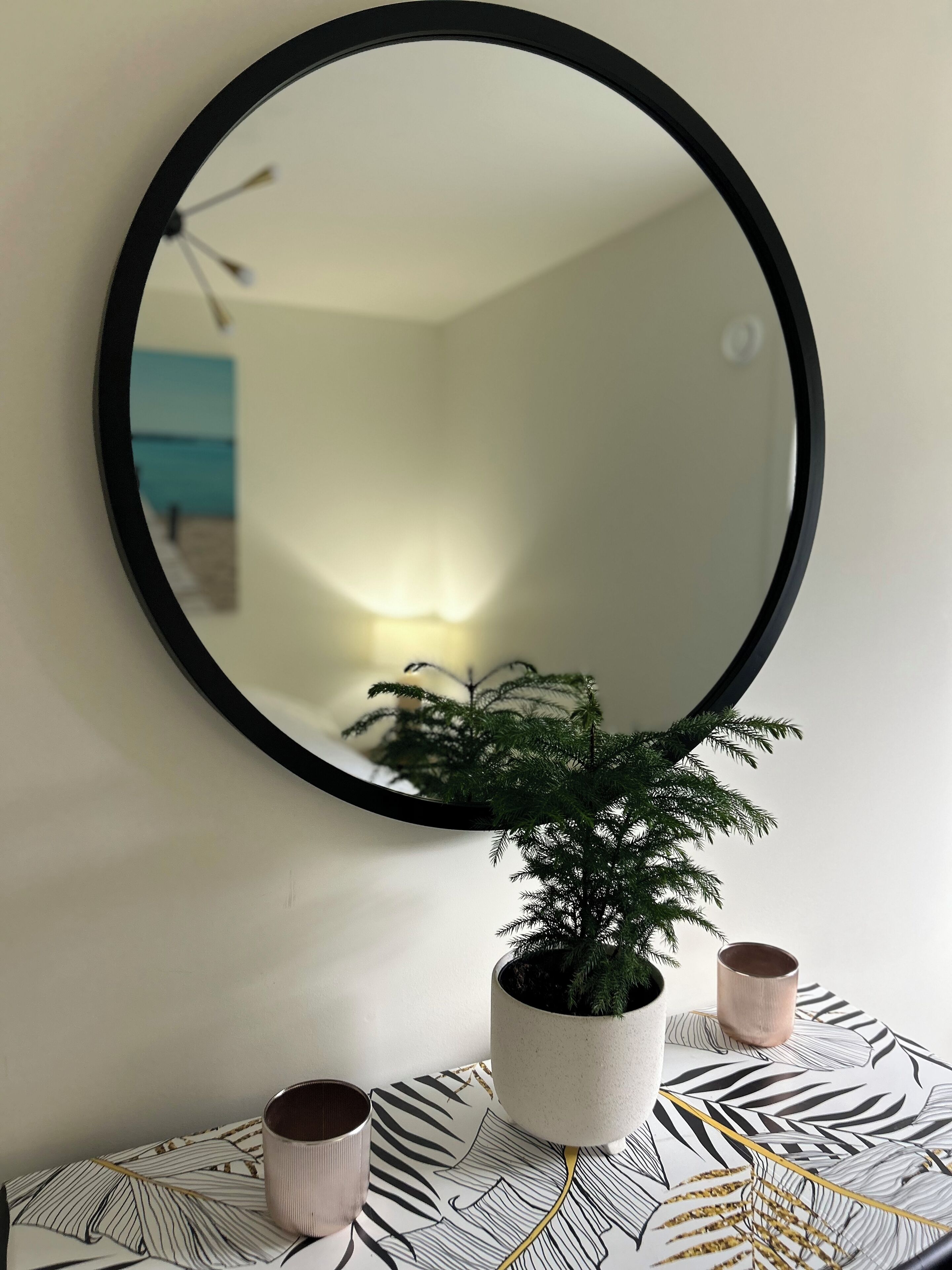 Ein runder Spiegel an der Wand, ein Blumentopf und zwei kleine Tassen auf einem gemusterten Tisch.