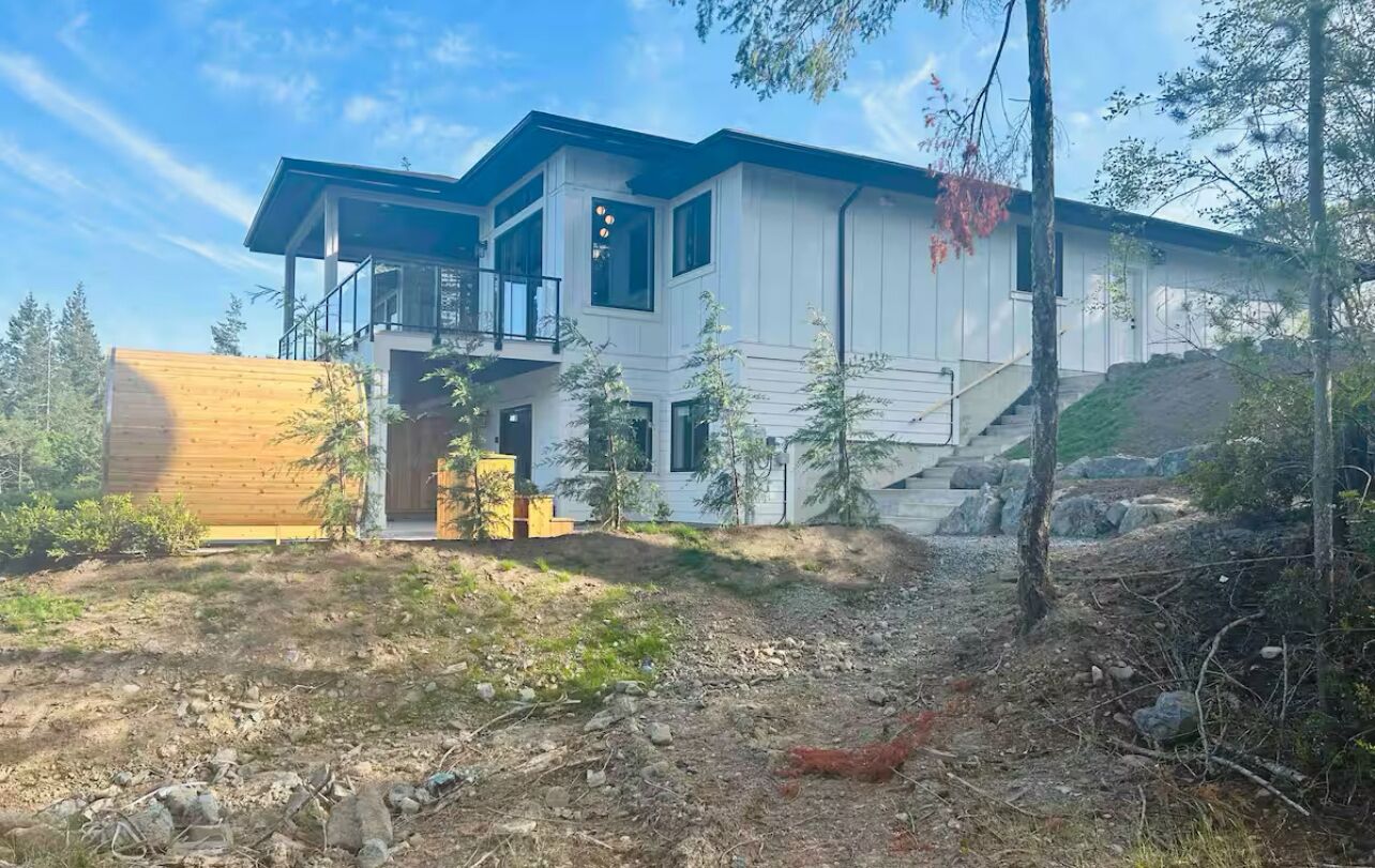 Una casa moderna de dos pisos con un amplio balcón, rodeada de árboles y un paisaje rocoso.