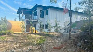 Una casa moderna de dos pisos con un amplio balcón, rodeada de árboles y un paisaje rocoso.