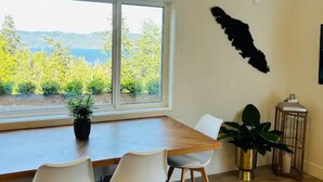 Un coin repas avec une table en bois et des chaises blanches, une plante en pot, et une vue sur les arbres et les montagnes par une grande fenêtre.