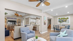 A spacious living room with a large round coffee table, two striped armchairs, and a ceiling fan. A kitchen with white cabinets and a dining area with a wooden table and chairs are visible in the background.