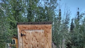A wooden structure with a door, surrounded by dense vegetation and a gravel path. - Alaskan Adventure Dry Cabin #2 (Fairbanks)
