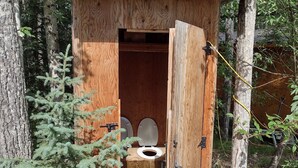 Een houten uitgebouw met een toilet en een deur, omgeven door bomen en groen.