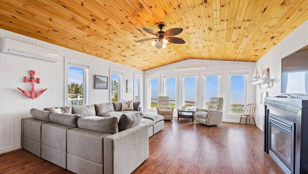 A spacious living room with a wooden ceiling, a large sectional sofa, a fireplace, and large windows offering a view of the outdoors.