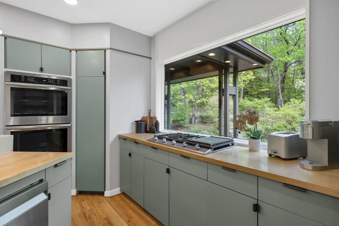 A modern kitchen with built-in appliances, a large window with a view of greenery, and a wooden countertop.
