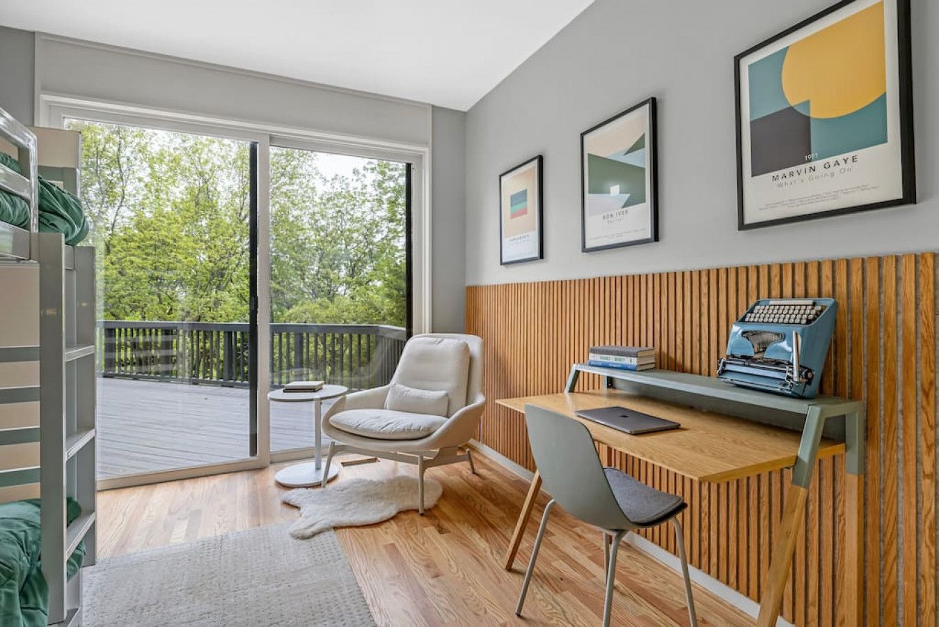A modern room with a wooden desk, a chair, a laptop, and a typewriter. There are framed pictures on the wall and a balcony with a view of trees.