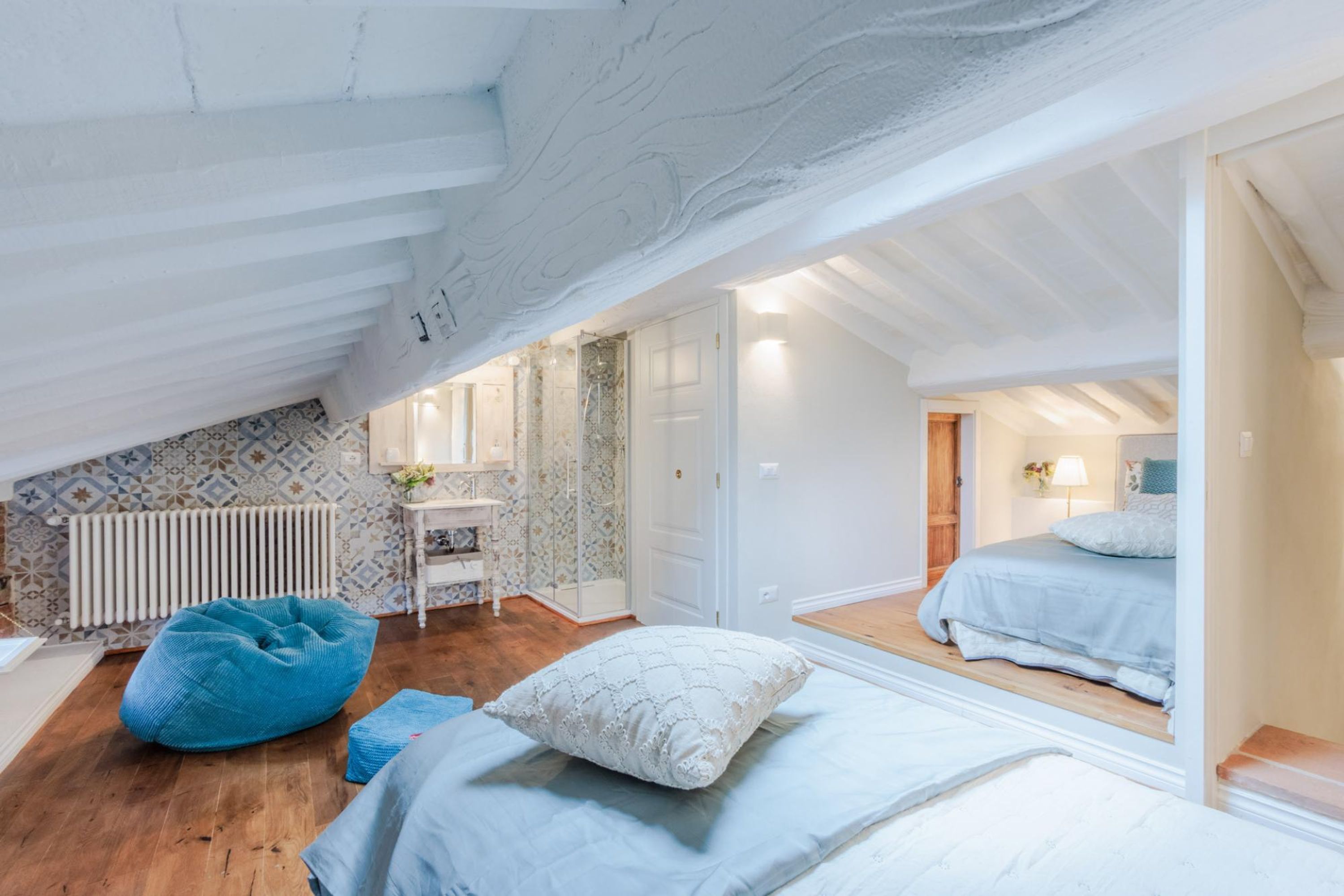 A bedroom with a sloped ceiling, a bed with a white duvet, a blue bean bag, a wooden floor, and a bathroom with a shower and a mirror.