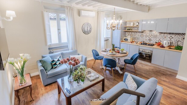 A modern living room with a grey sofa, a coffee table, and a dining area with blue chairs. There is a kitchen with grey cabinets and a tiled backsplash. - Casa Chiostro in Lucca (Lucca)