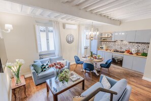 A modern living room with a grey sofa, a coffee table, and a dining area with blue chairs. There is a kitchen with grey cabinets and a tiled backsplash. - Casa Chiostro in Lucca (Lucca)