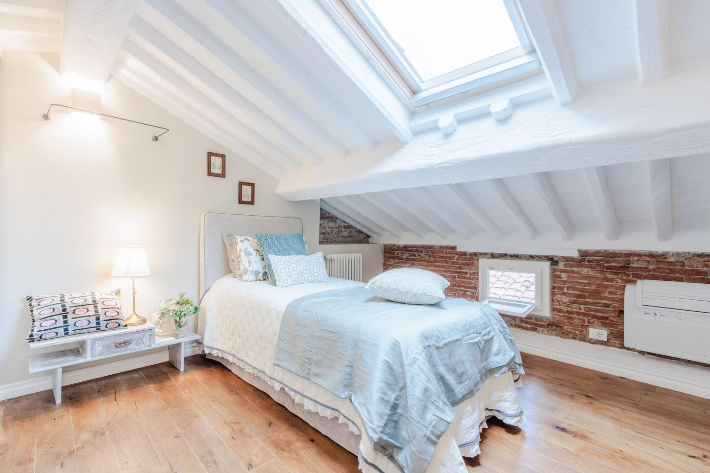A bedroom with a skylight, wooden flooring, a bed with a blue bedspread, a nightstand with a lamp, and a brick wall.
