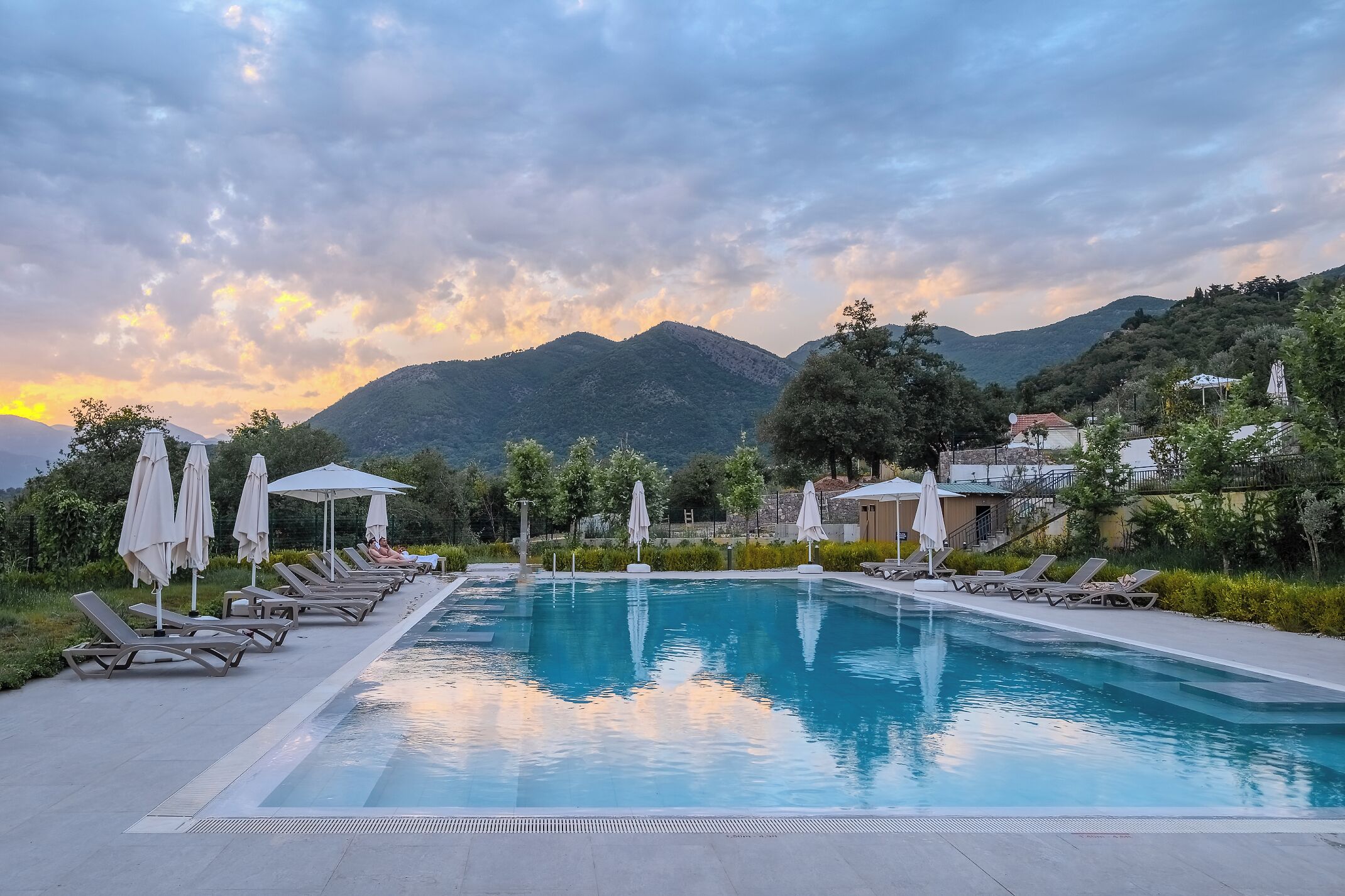 A swimming pool with lounge chairs and umbrellas, surrounded by greenery and mountains in the background.