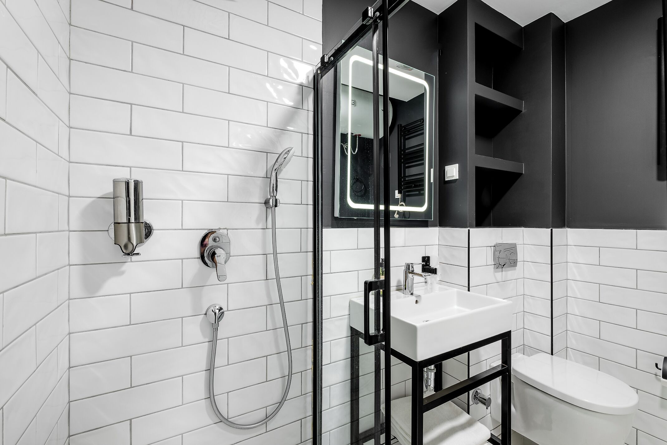 Un baño moderno con paredes de azulejos blancos, un lavamanos con marco negro, un inodoro y una ducha con puerta de vidrio.