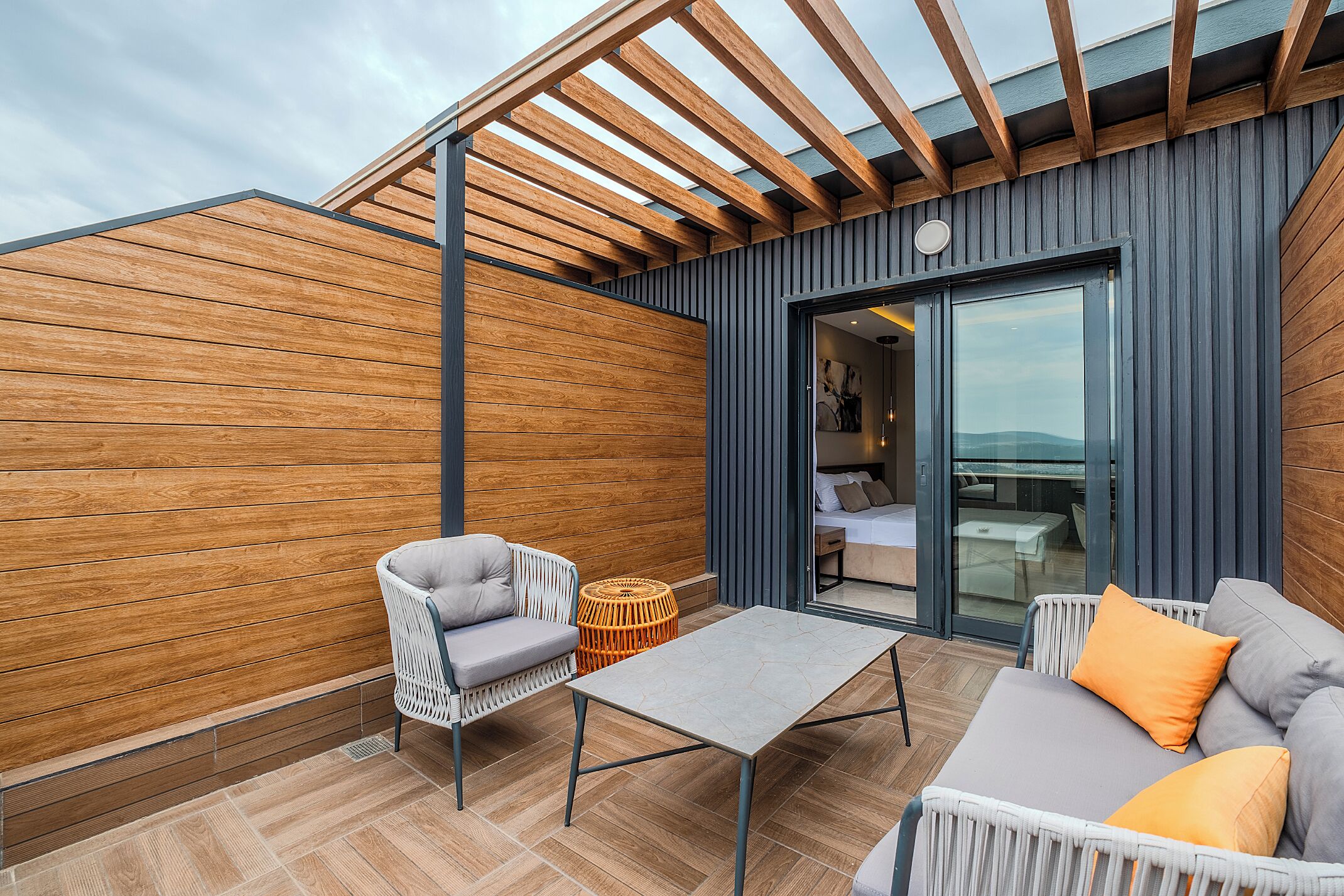 Un patio exterior moderno con una pérgola de madera, un sofá gris, una silla de mimbre blanca y una mesita.