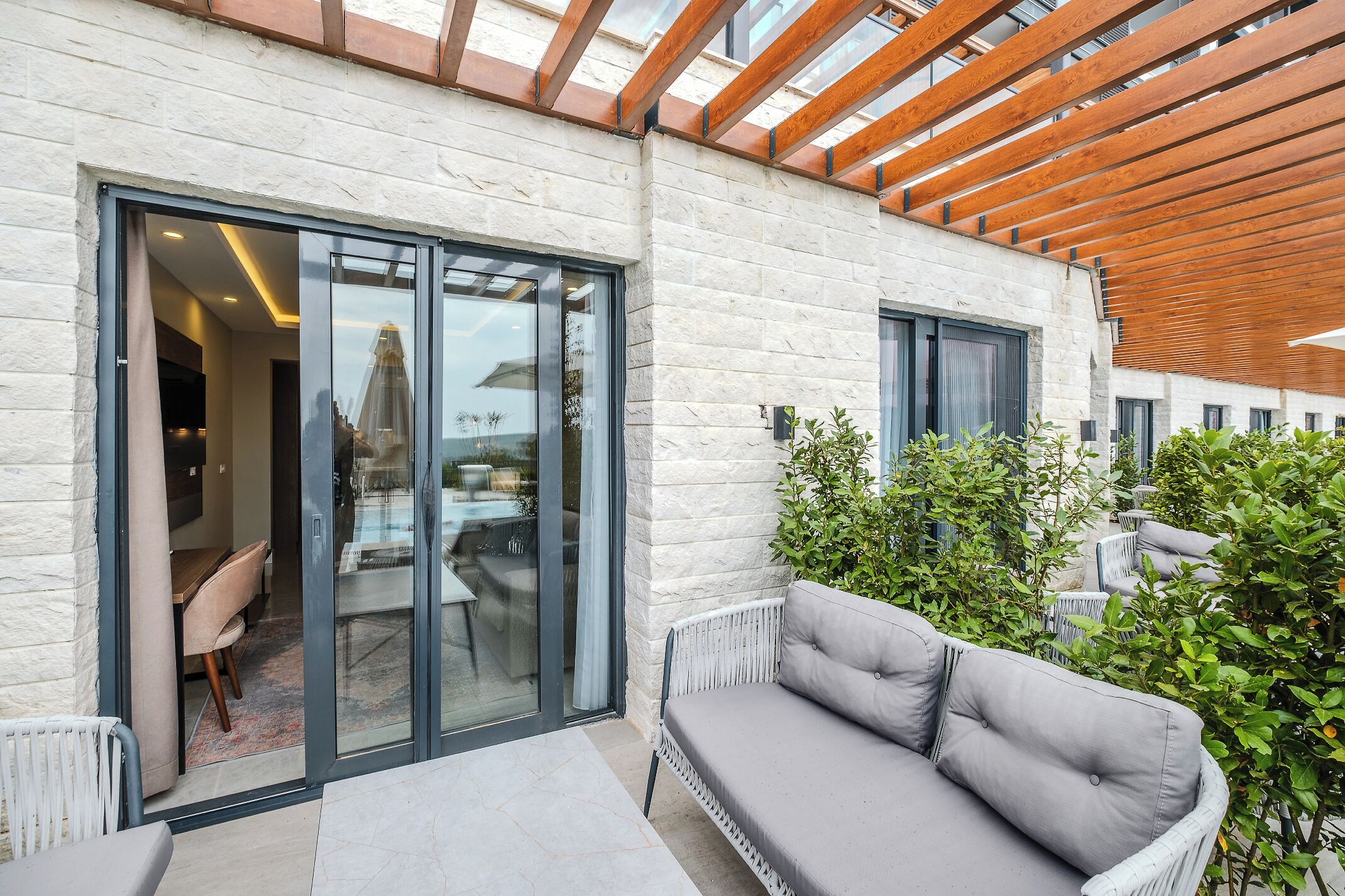 A modern building with a glass door entrance, a grey cushioned outdoor seating area, and a wooden pergola.