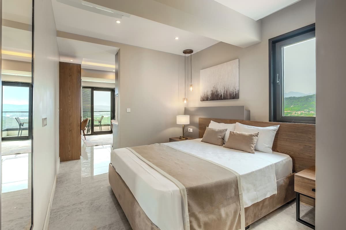 A modern bedroom with a large bed, a nightstand, a lamp, and a view of the outdoors.