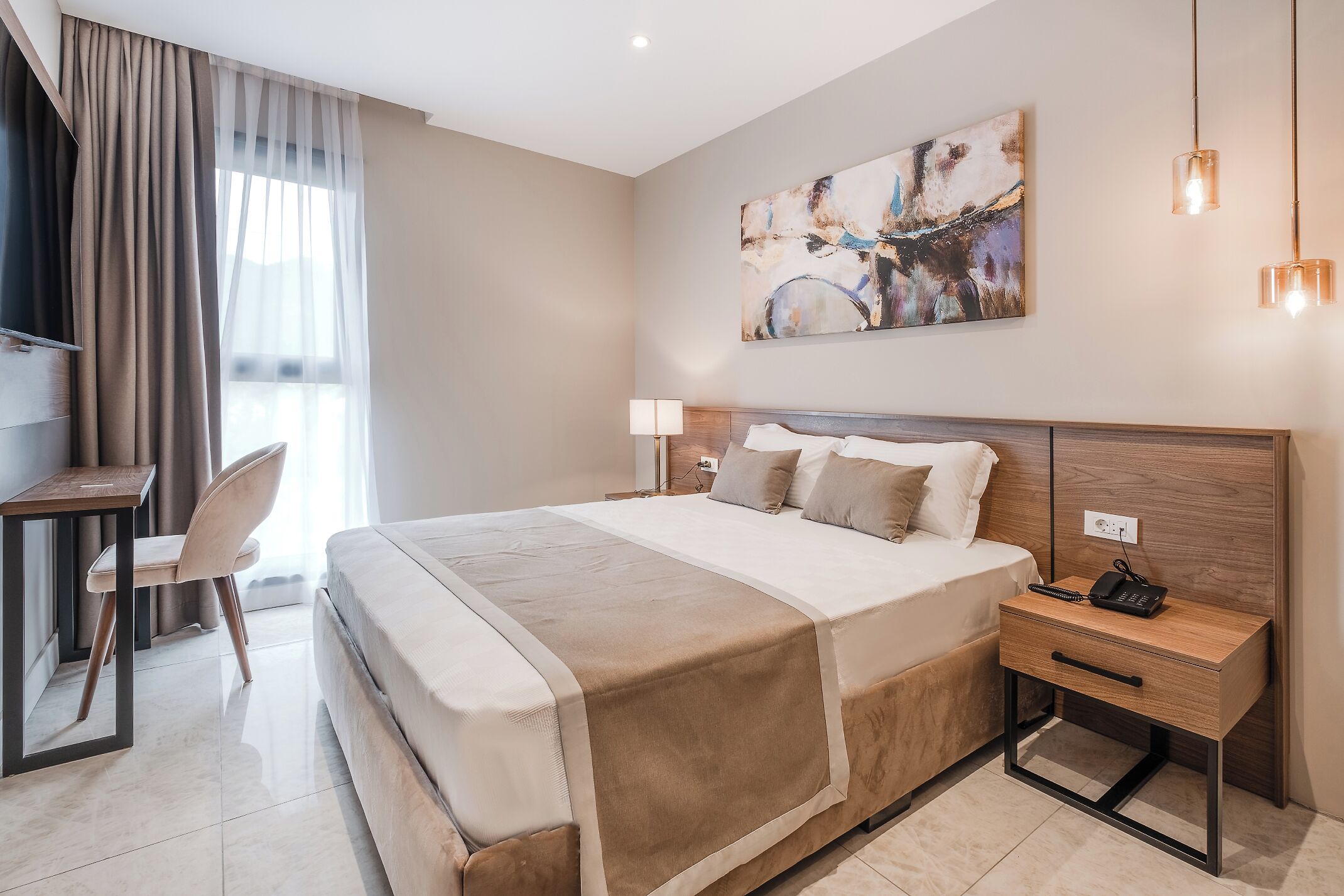 A modern hotel room with a large bed, bedside table, and a desk with a chair.