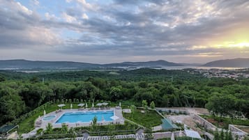 Un complexe hôtelier doté d’une grande piscine entourée de chaises longues et de parasols, le tout situé dans un cadre verdoyant luxuriant, avec des montagnes qui se dessinent à l’horizon.