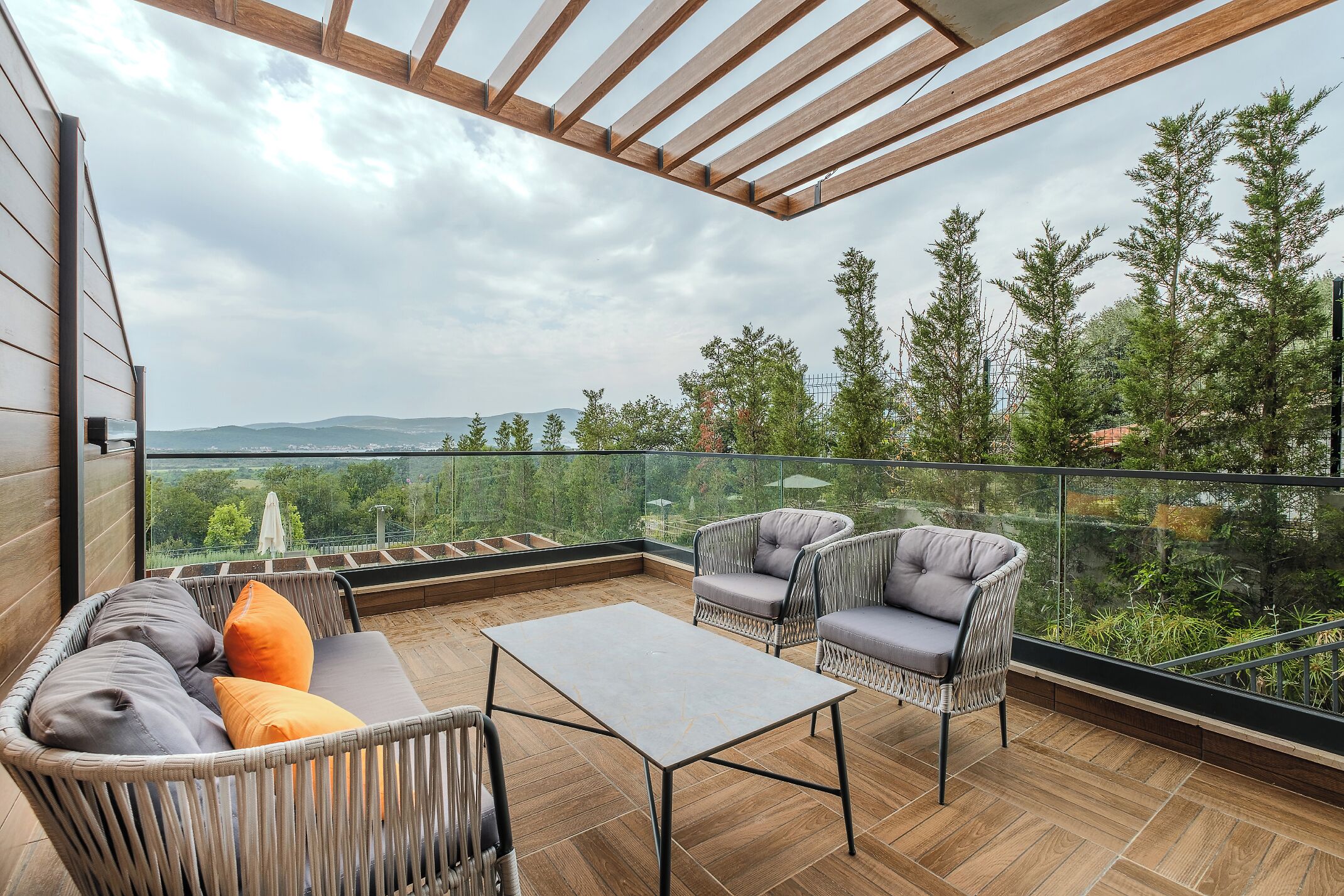 A balcony with wicker furniture, a table, and a glass railing overlooking a forested area.