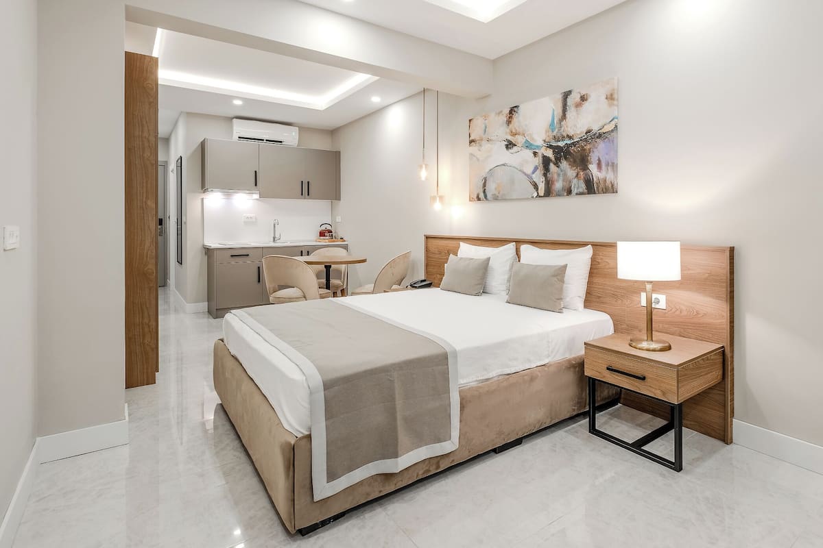 A modern hotel room with a large bed, bedside table, and a view into the kitchen area.