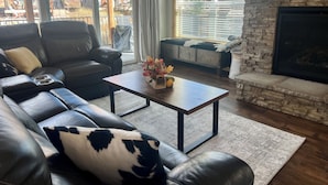 A living room with a leather sofa, a coffee table with flowers, a stone fireplace, and large windows with blinds. - 2 Kg Beds+ Walk to Ponds, Pool, Hot tub&River. Short Drive to RMNP & Winter Park (Granby)