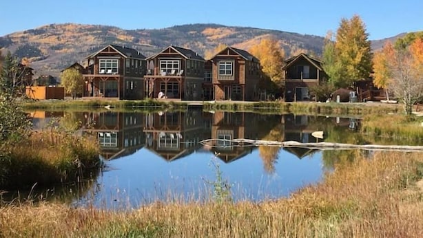 A row of wooden cabins with a reflective pond in front, set against a backdrop of autumn-colored trees and hills. - 2 Kg Beds+ Walk to Ponds, Pool, Hot tub&River. Short Drive to RMNP & Winter Park (Granby)