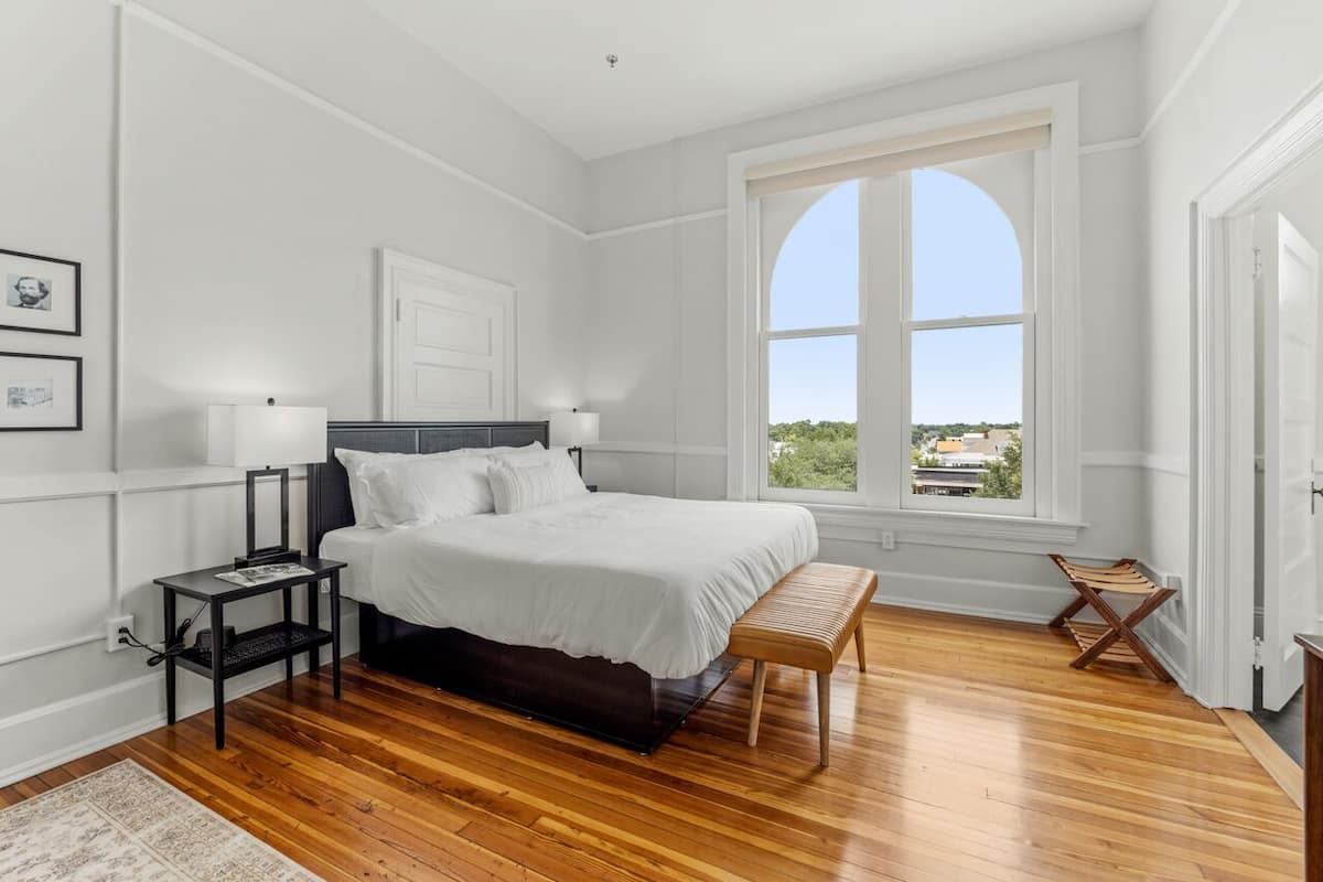 A bedroom with a large bed, a nightstand, a bench, and a window with a view.