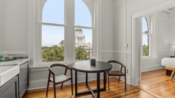 A room with a large arched window, a round table with chairs, a bed with a white headboard, and a view of a building with a dome. - The McKey (Valdosta)