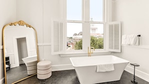A modern bathroom with a freestanding bathtub, a large mirror with an ornate frame, and a view of a cityscape through large windows. - The McKey (Valdosta)