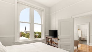 A bedroom with a large window, a bed, a TV on a wooden stand, and a door leading to another room. - The McKey (Valdosta)