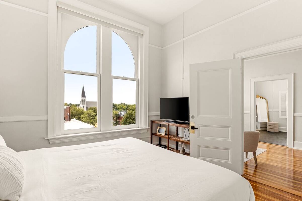 A bedroom with a large window, a bed, a TV on a wooden stand, and a door leading to another room.