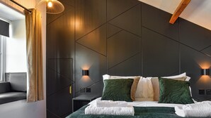 A modern bedroom with a dark wall panel design, a bed with green and white bedding, two wall-mounted reading lights, and a window with curtains.