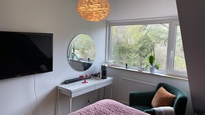 Un dormitorio con cama, escritorio, silla, televisión, ventana con vistas a árboles y una lámpara colgante.