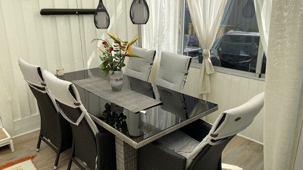 Una mesa de comedor con cubierta de vidrio rodeada de sillas de mimbre, un florero con flores y una pared blanca con ventana y cortinas.
